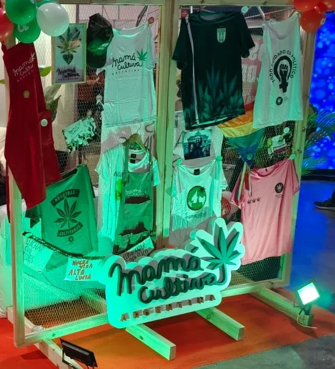 Stand de Mamá Cultiva Argentina con remeras colgadas y el cartel de la organización